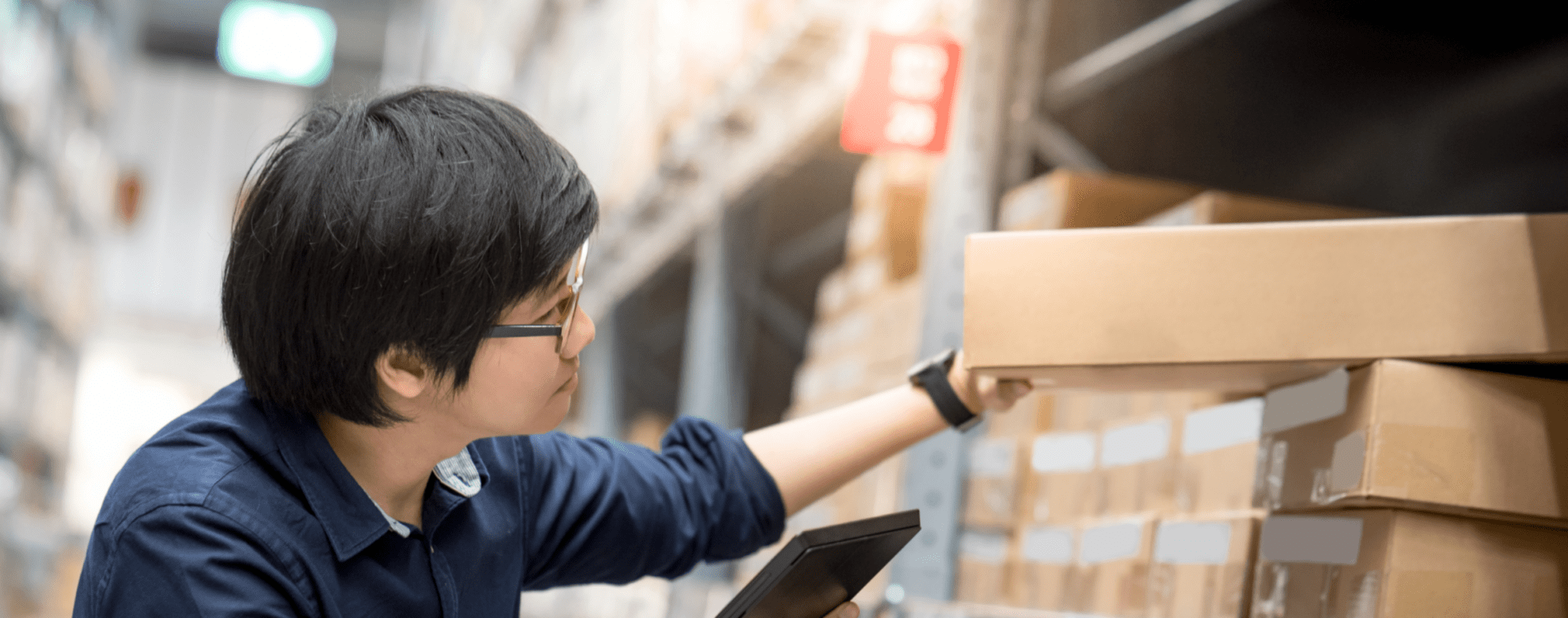 Young Asian man doing stocktaking of product in cardboard box on shelves in warehouse by using digital tablet