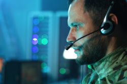 Pan around view of serious commander in camouflage speaking on headset and using computer while working in mission control center during war