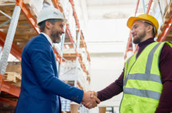 Waist up portrait of handsome mature businessman shaking hands with worker wearing hardhat standing against warehouse shelves in background