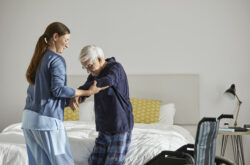 Caregiver supporting senior man to stand. Mid adult female nurse is assisting elderly male at home. They are in bedroom.