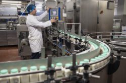 Three-quarter length side view of man in white coat and hair net controlling movement of containers with green caps processing in single file on conveyor belt. / Female Focus Collection